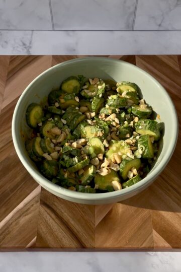 Bowl of cucumber salad with chopped peanuts and herbs on a wooden table.