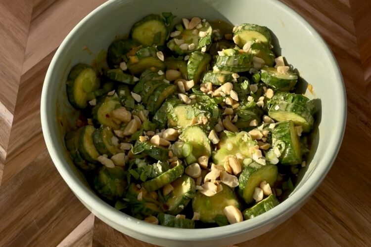 Bowl of cucumber salad with chopped peanuts and herbs on a wooden table.